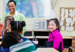 An image of an Asian American girl smiling at the camera in a diverse classroom. Credit: Canva.